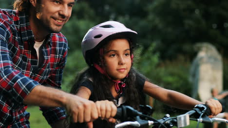 Closeup.-Portrait-of-a-little-girl.-Her-dad-is-teaching-her-to-ride-a-bike.-He-supports-her.-Moving-camera.-Blurred-background